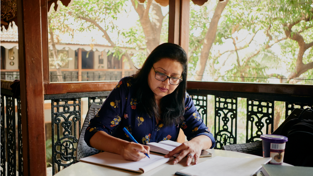 Eine Gastgeberin mit Brille und einem Hemd mit Blumenmuster schreibt etwas auf einen Block Papier, während sie auf einer Veranda in Mumbai, Indien, sitzt.