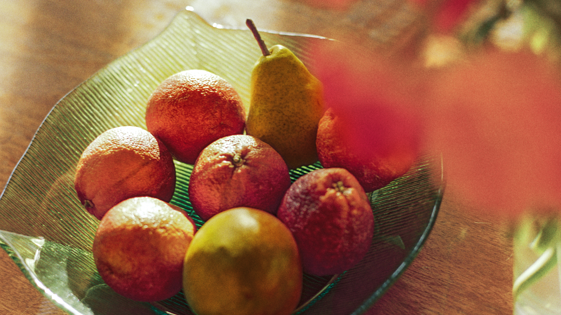 Tá torthaí úra, citris agus piorraí, i mbabhla glas ar bhord adhmaid in aice le vása bláthanna.