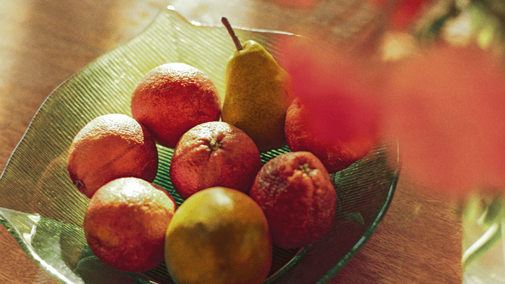 Tá torthaí úra, citris agus piorraí, i mbabhla glas ar bhord adhmaid in aice le vása bláthanna.