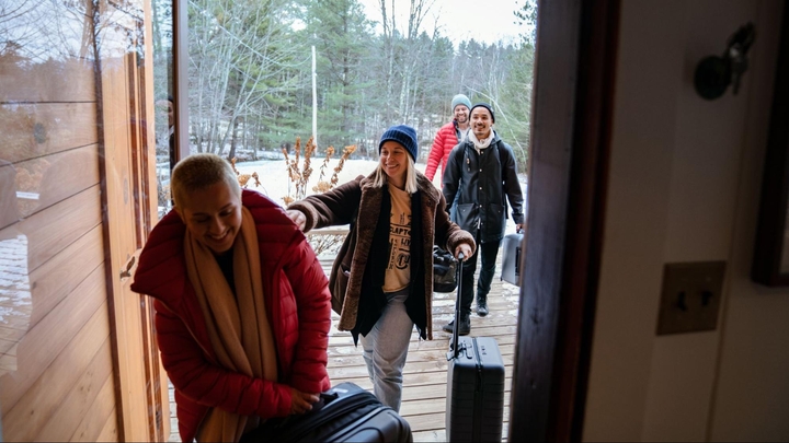 Four people wearing coats smile as they carry suitcases from a snowy porch through the open door of a house.