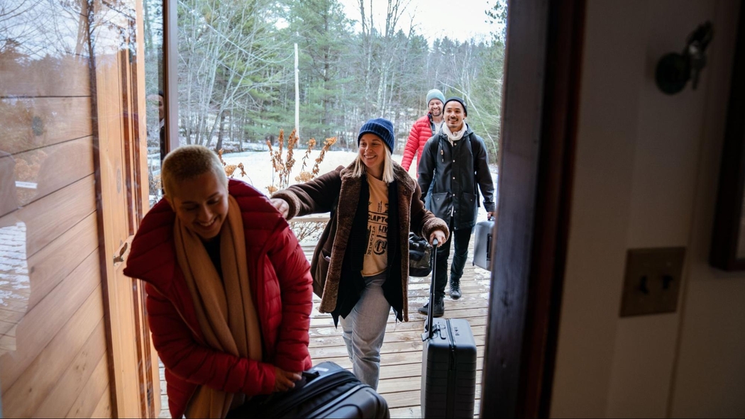 Cuatro personas con abrigos sonríen mientras llevan valijas desde una galería cubierta de nieve a través de la puerta abierta de una casa.