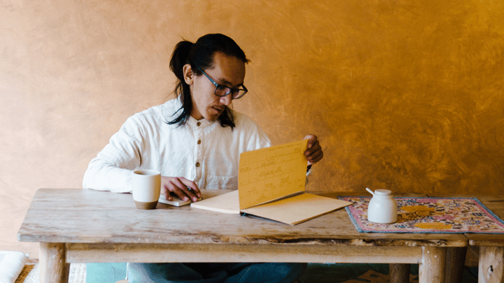 A person sitting cross-legged on the floor reviews a notebook that sits on a low wooden table.