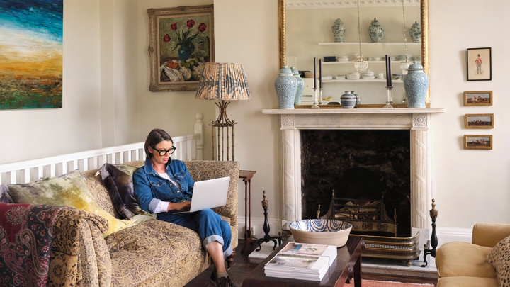 Tummatukkaisella henkilöllä on yllään farkkutakki ja farkut. Hän istuu vaaleanruskealla sohvalla takan vieressä, ja hänellä on sylissään kannettava tietokone.