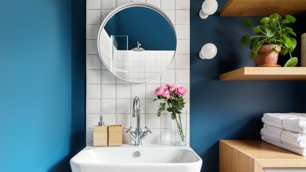 Una toletta da bagno pulita e moderna con specchio rotondo, alzatina in piastrelle bianche e dettagli in legno di quercia.