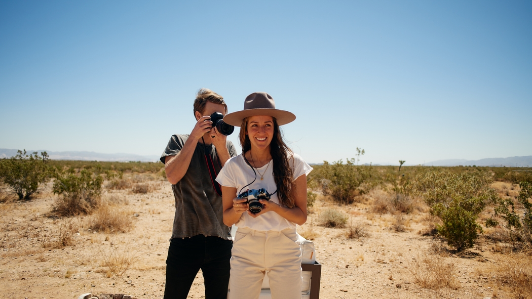 Two people stand, one in front of the other, in a desert landscape under a clear blue sky. Each is holding a camera.