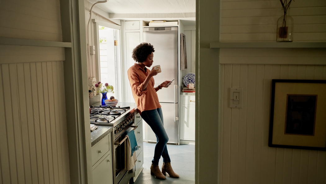 Uma pessoa usando jeans e uma camisa laranja está em pé em uma cozinha segurando uma caneca e olhando para o celular.