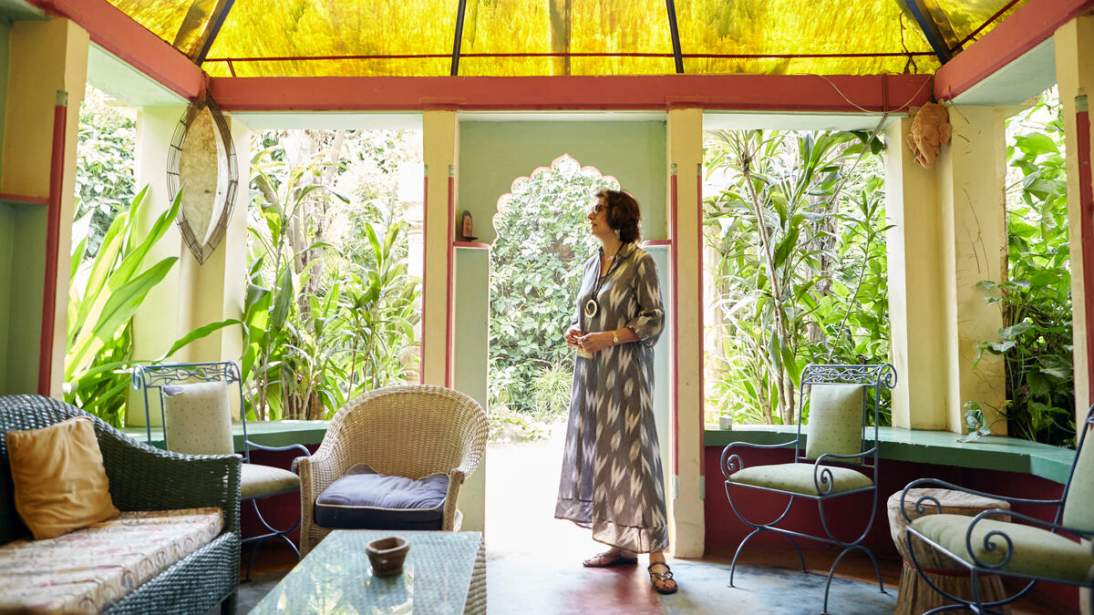 Kona í gráum kjól með hvítu mynstri stendur á verönd með gulu þaki umkringd plöntum.