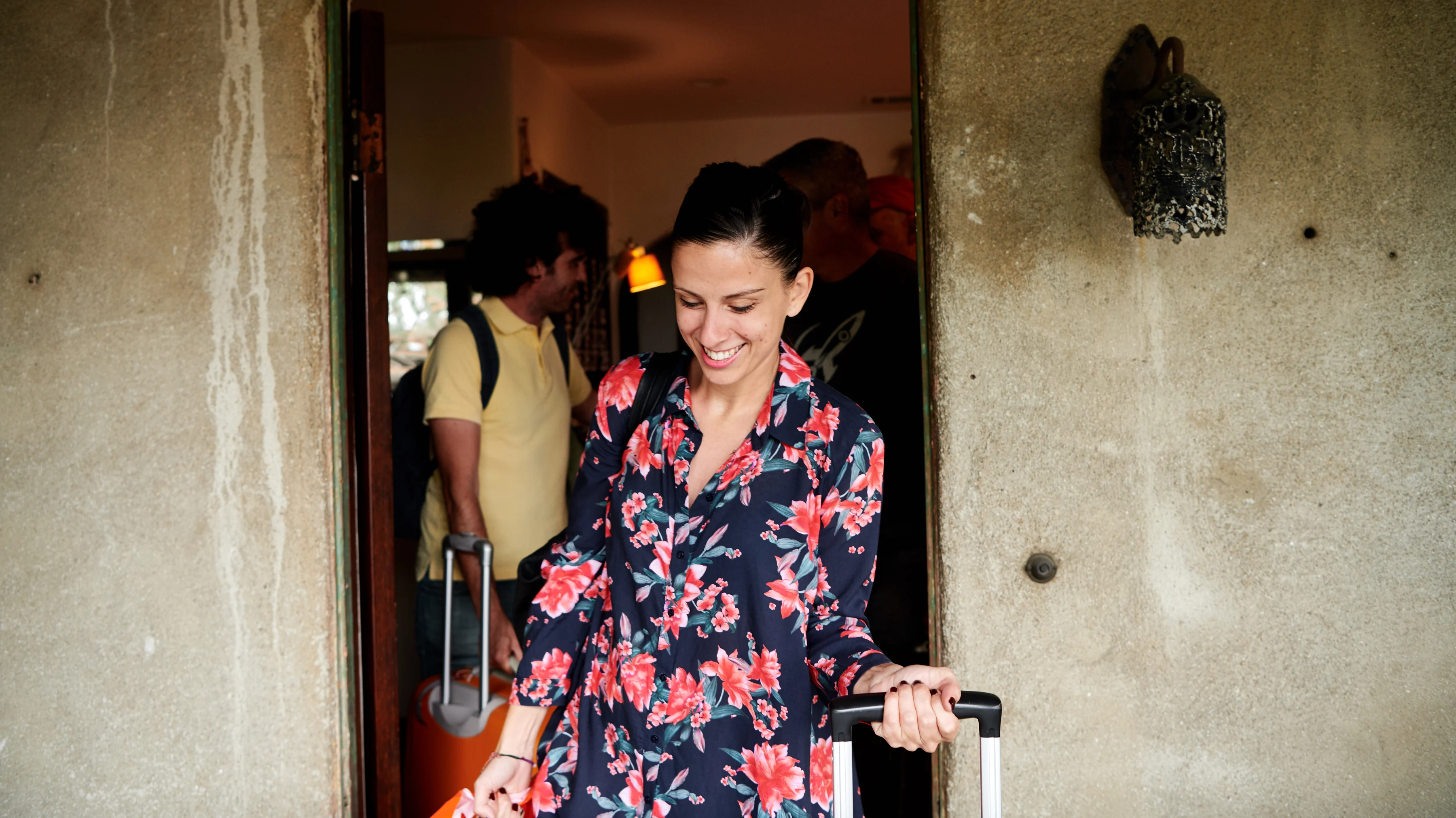 Woman smiling and holding luggage, walking through a door followed by other people with luggage.