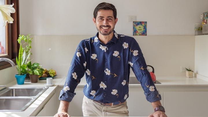 A man wearing a dark blue button-up shirt with a white flower print smiles with his hands on a white kitchen countertop.