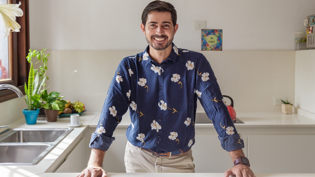 A man wearing a dark blue button-up shirt with a white flower print smiles with his hands on a white kitchen countertop.