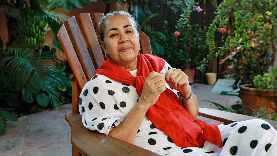 A woman sits in a wooden chair wearing a polka dot dress and red scarf.