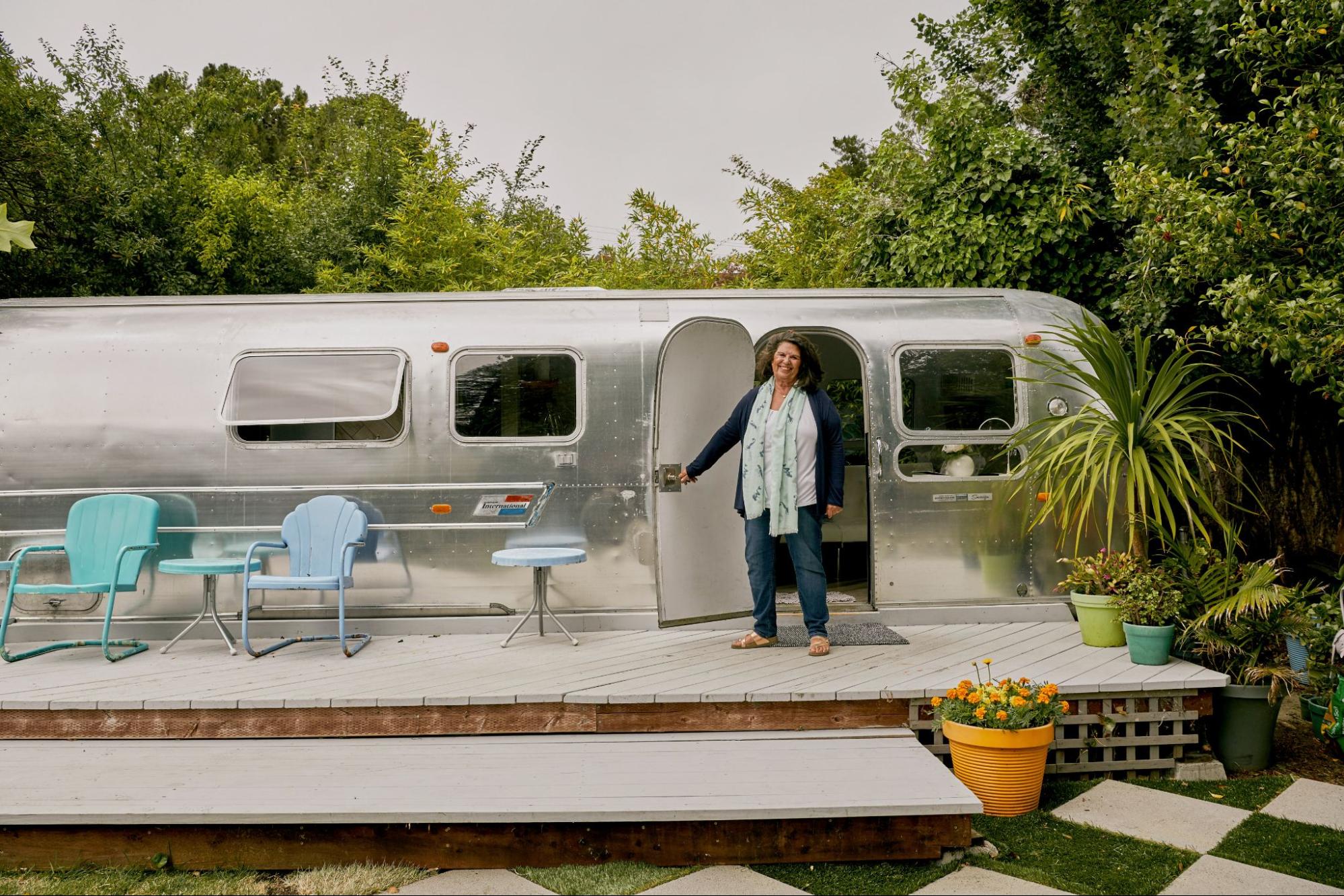 Un anfitrión está de pie en un deck de madera y manteien abierta la puerta de una casa rodante, un ejemplo de un  tipo de propiedad que es un alojamiento único.