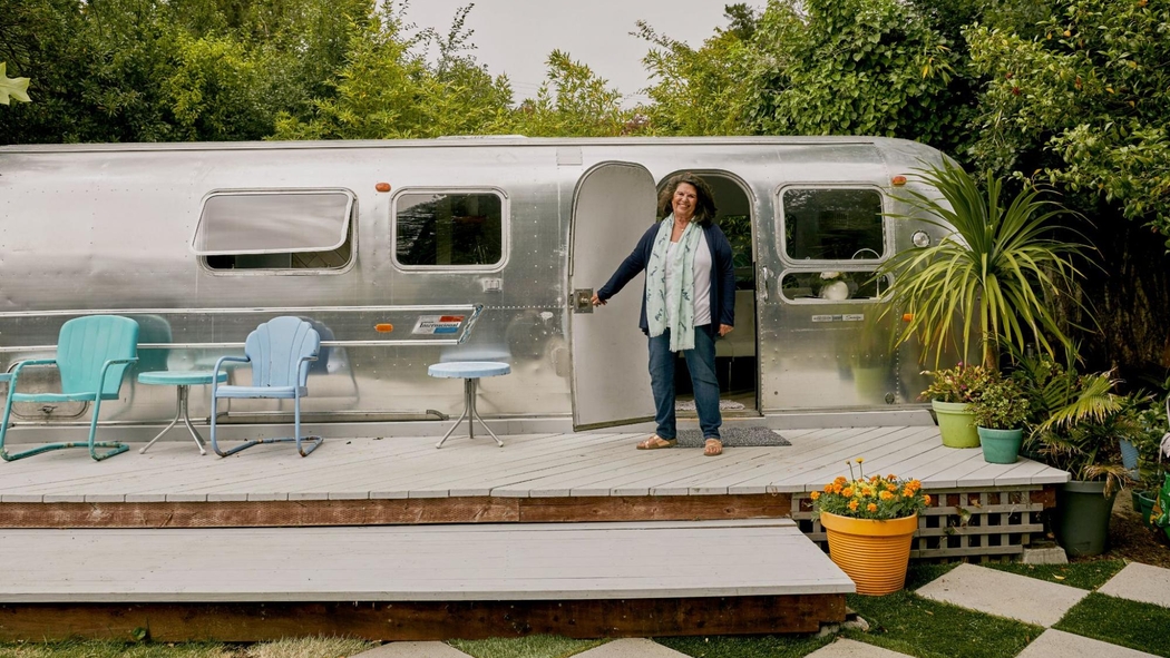 Un hôte se tient sur une terrasse en bois et maintient ouverte la porte d'un logement unique : une caravane Airstream.