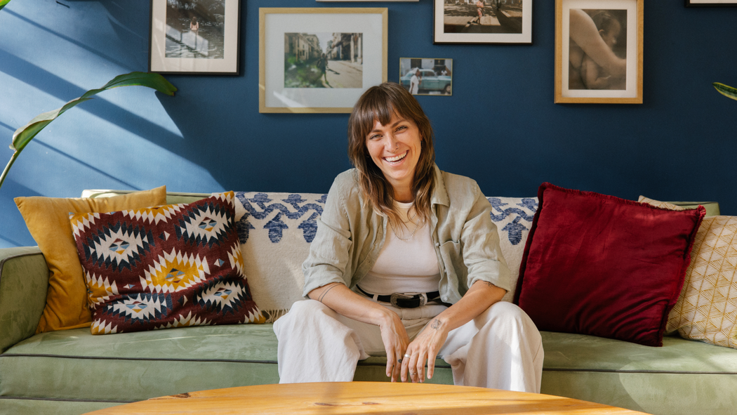 Una anfitriona en Airbnb sonríe y está sentada en un sillón de felpa con almohadones de colores. Detrás hay una pared adornada con fotos de personas.