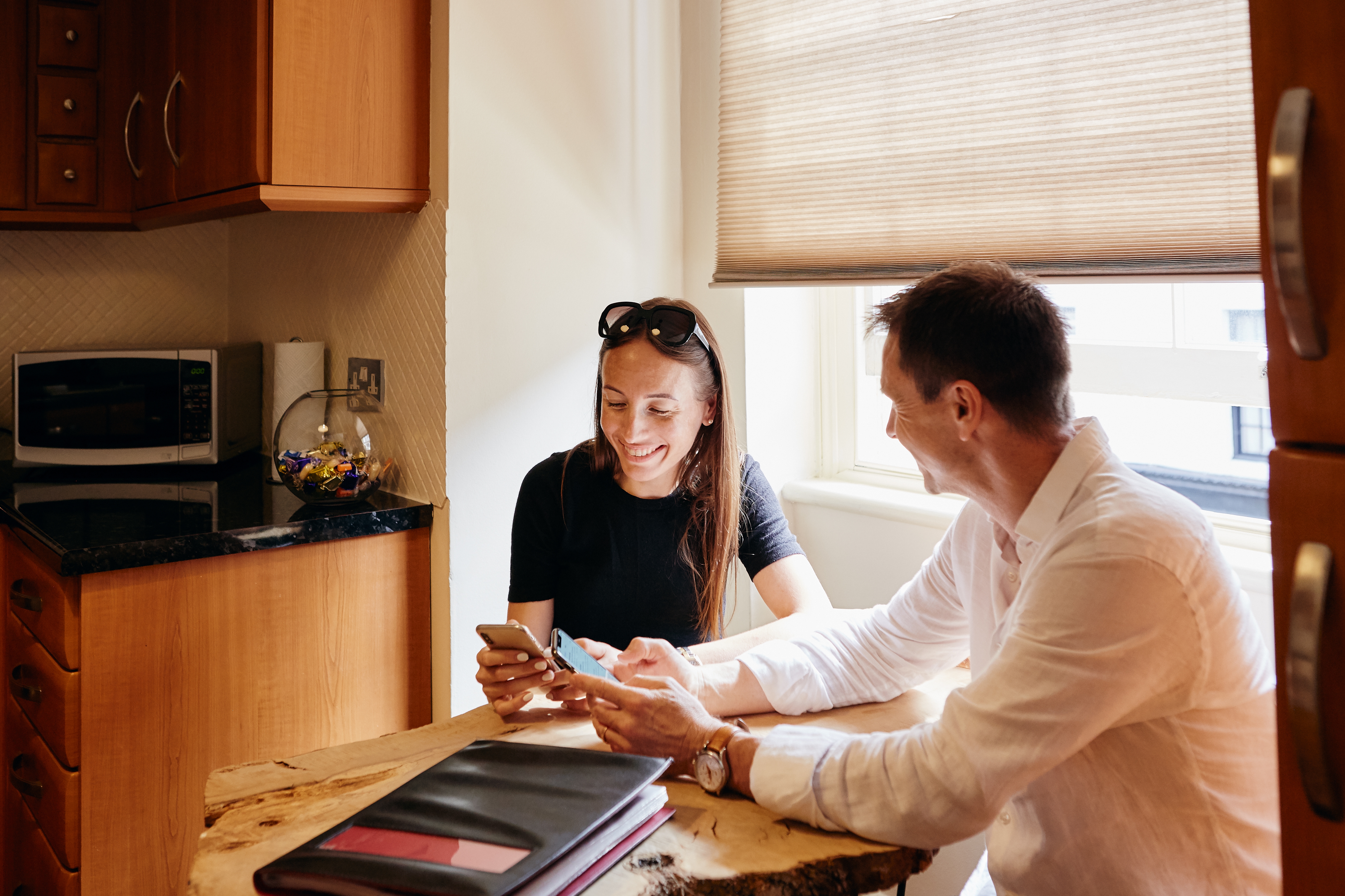 Dues persones assegudes a una taula de cuina petita feta amb un tronc de fusta somriuen mentre fan servir el mòbil.