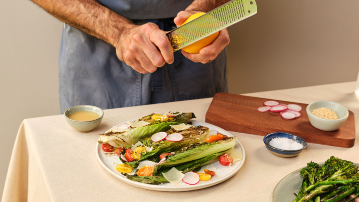 Eine Person verwendet eine Raspel, um Zitronenschale zu einem Salat aus Römersalatblättern, in Scheiben geschnittenen roten Radieschen und Kirschtomaten hinzuzufügen.