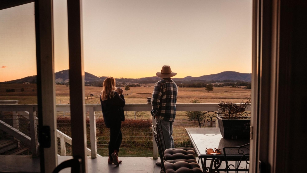 Two people holding mugs look at the mountain-lined horizon from an elevated porch.