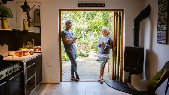 İki nəfər geniş qapı girişində dayanıb söhbət edir, arxa fonda yaşıllıq, ön planda isə odun sobası olan mətbəx var.