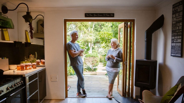 Two people talk in a wide doorway, with greenery in the background and a kitchen with a woodstove in the foreground.