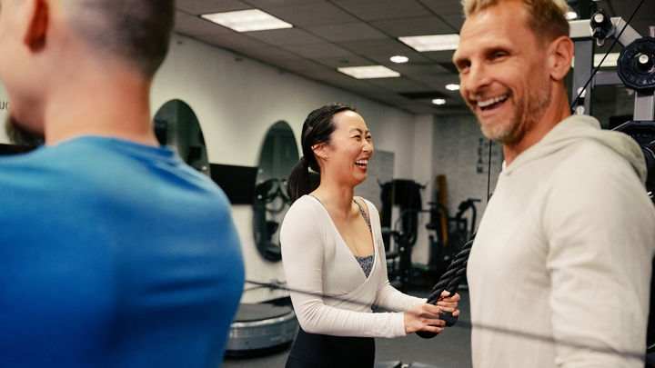 Duas pessoas sorriem em uma academia repleta de espelhos e equipamentos de treino.