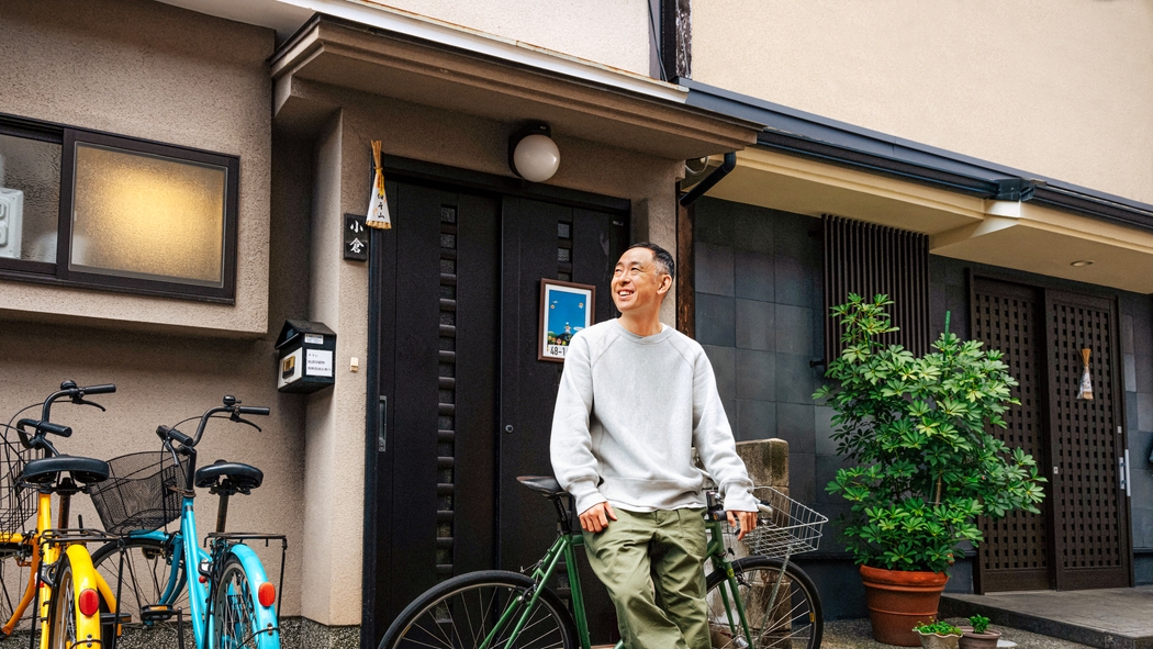 شخص يرتدي سترة رمادية وسروالًا أخضر اللون يستند إلى دراجة أمام باب أسود ونباتات مزروعة في أوعية.