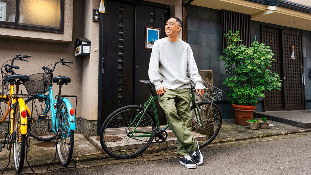 A person wearing a gray sweatshirt and green pants leans against a bicycle in front of a black door and potted plants.