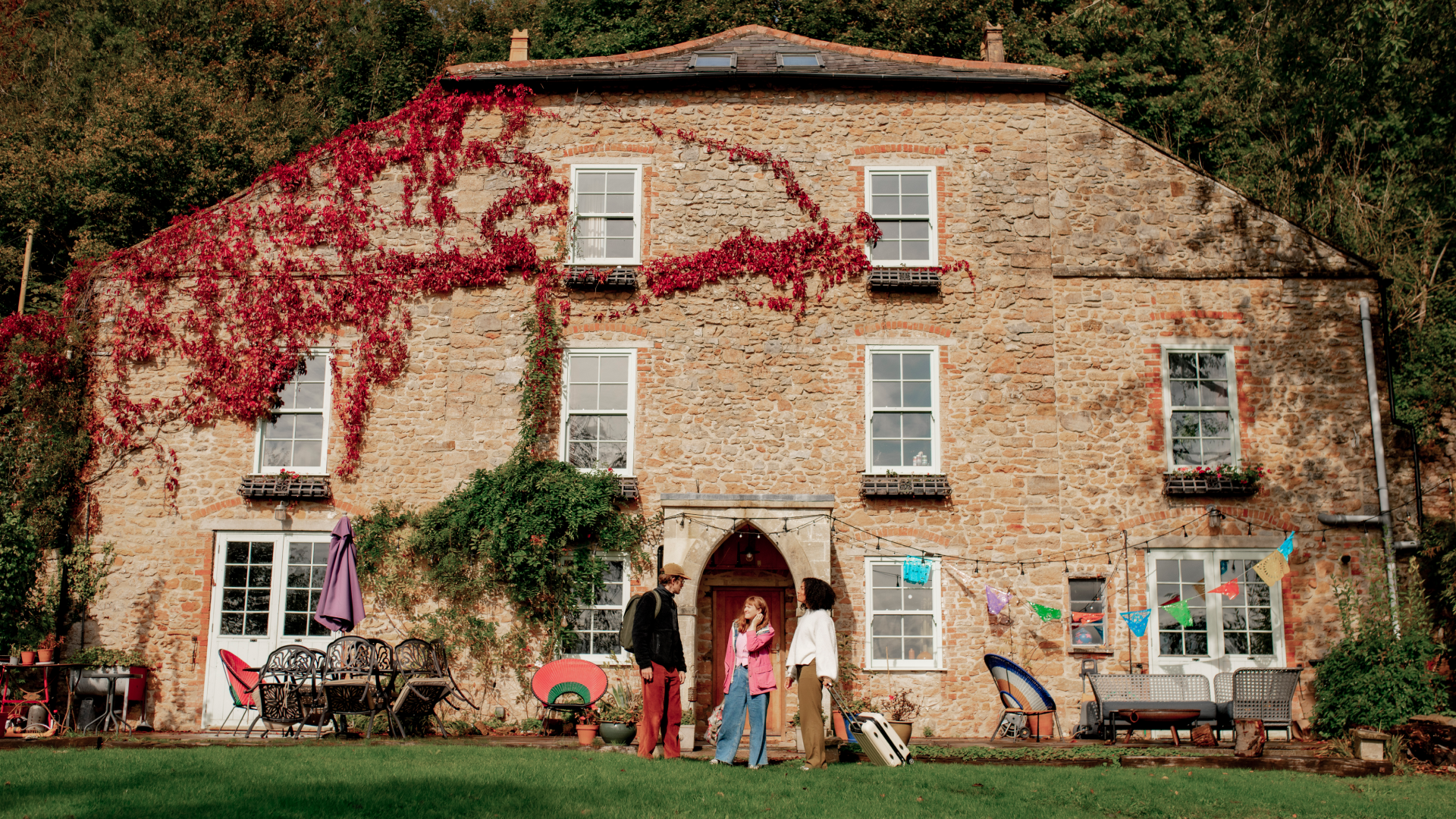 Watu watatu wamesimama na kuzungumza katika mwanga wa jua nje ya jengo la mawe la zamani lililofunikwa kwa mwefeu mwekundu na wa kijani.