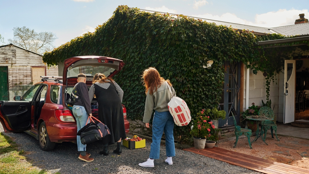 Drie mensen pakken koffers uit de kofferbak van een rode auto bij een woning bedekt met klimop.