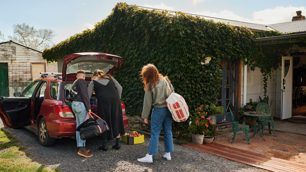 Three people unpack bags from the trunk of a red car outside a house covered in ivy.