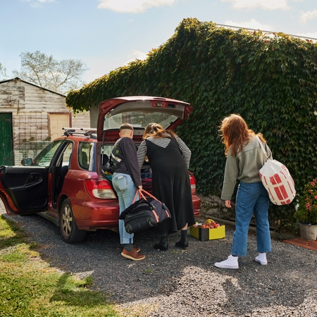 Three people unpack bags from the trunk of a red car outside a house covered in ivy.