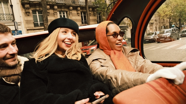 Trīs mēteļos un šallēs tērpti cilvēki sēž aizmugurējā sēdeklī braucošā brīvdabas transportlīdzeklī, kas šķērso pilsētas ielu.