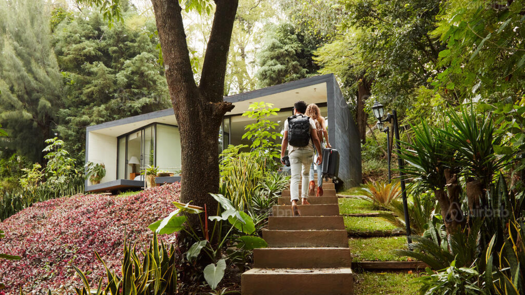 Deux personnes avec des valises arrivent dans une maison. Ils montent un escalier extérieur, entouré d'arbres et de verdure.