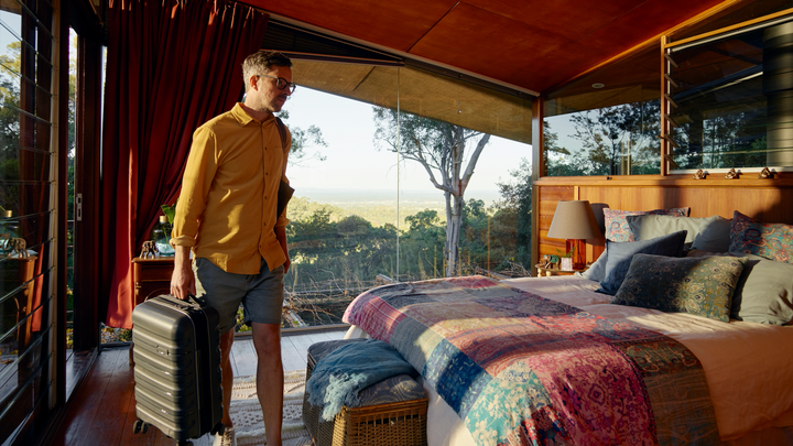 A person carries a suitcase in a large bedroom with windows facing greenery.