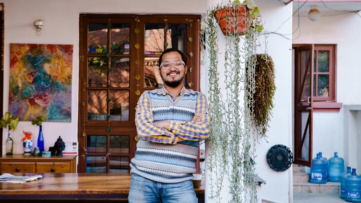 Une personne souriante, vêtue d'un pull sans manches rayé bleu et blanc et d'une chemise à carreaux, est appuyée contre un mur blanc couvert de plantes.