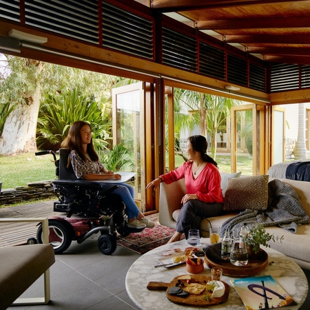 Two women sit in an airy room having a conversation. One woman is using a wheelchair, and the other is seated on a couch.