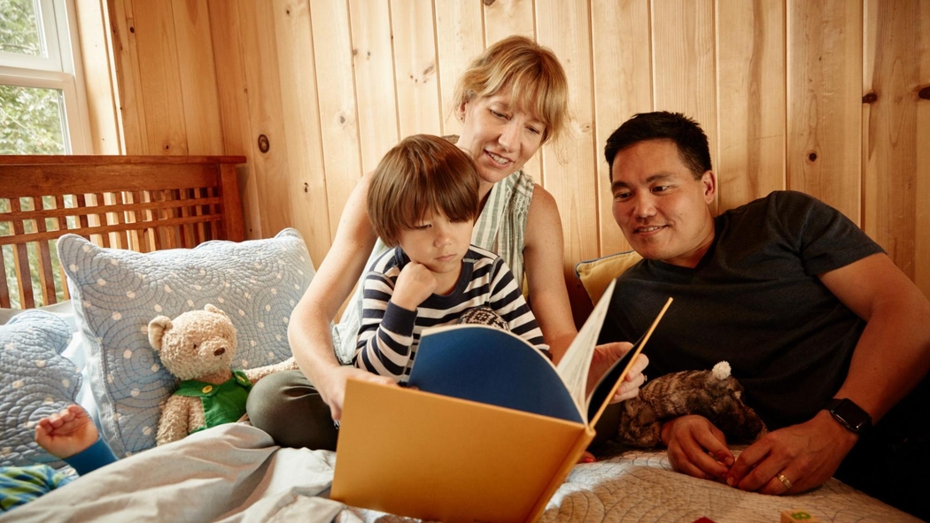 Two adults read a book to a toddler as they lounge on a bed. A sibling and a teddy bear are tucked in under a duvet.