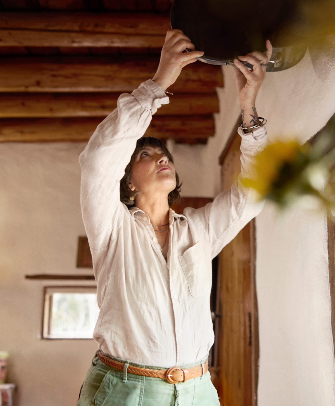Superhost Sadie räumt einen Kochtopf aus dem Weg, indem sie ihn an die Decke hängt. Im Hintergrund leuchtet eine Stehlampe neben einem Korbstuhl.
