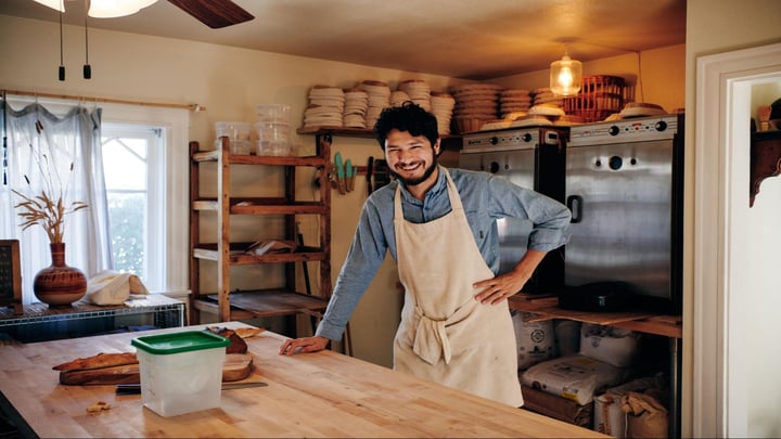 Seseorang yang mengenakan celemek berdiri di dekat kitchen-island dengan roti baguette terletak di atas talenan di samping pisau bergerigi.
