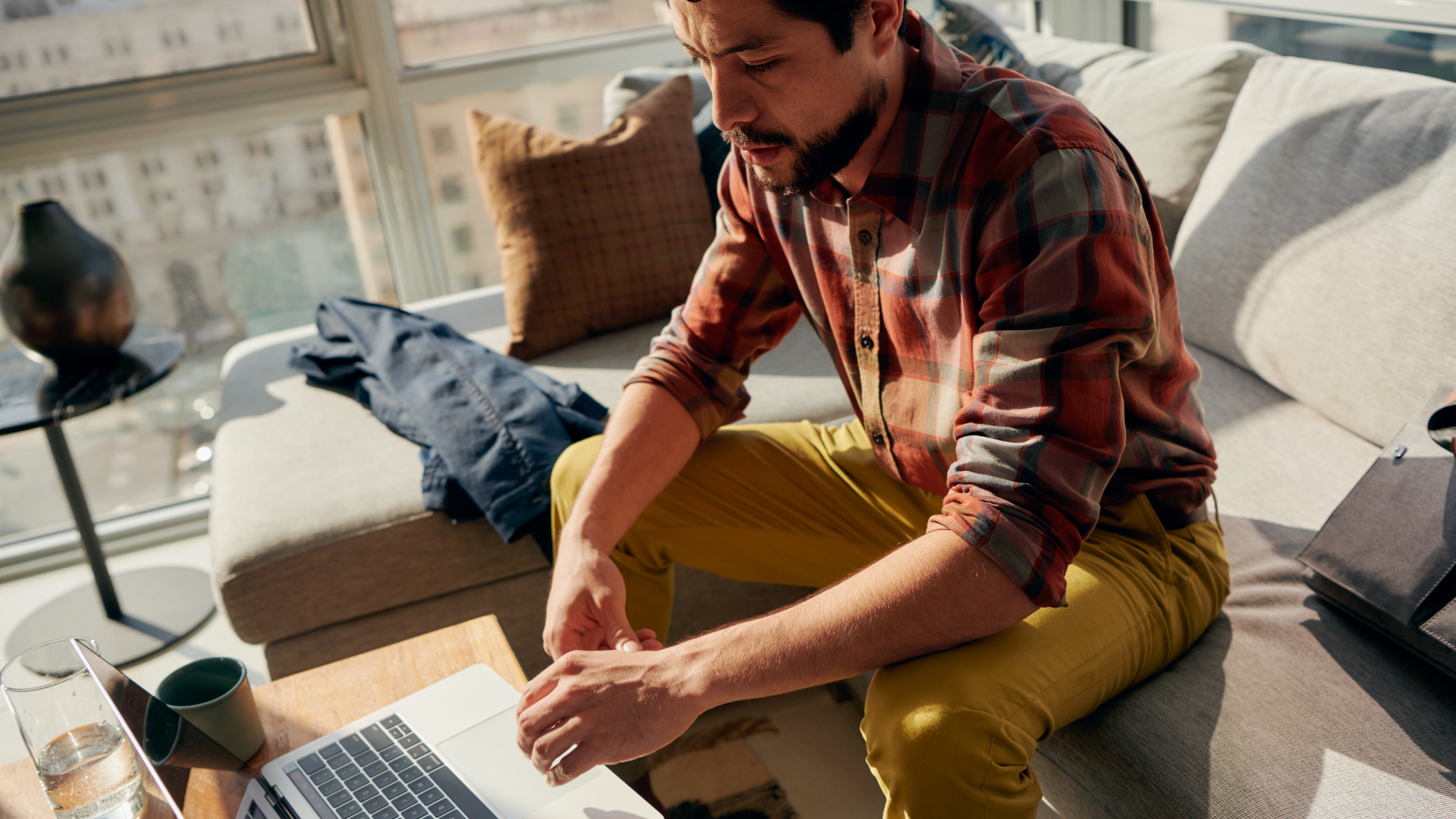 एक व्यक्ति काउच पर बैठा हुआ है और उसके सामने मौजूद कॉफ़ी टेबल पर एक लैपटॉप रखा हुआ है। खिड़की से धूप की रोशनी अंदर आ रही है।
