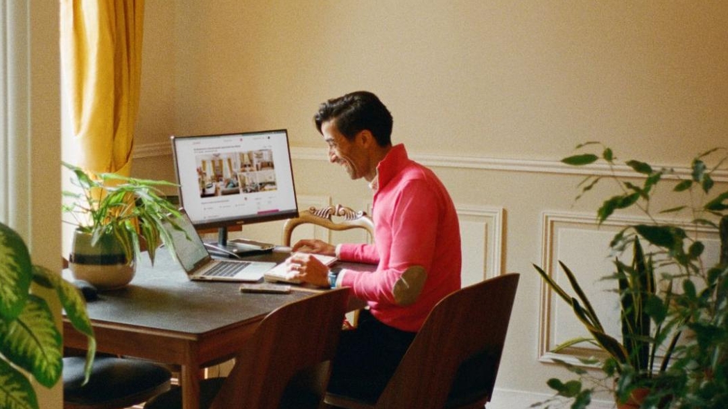 Человек сидит за рабочим столом с открытым ноутбуком и монитором перед ним.