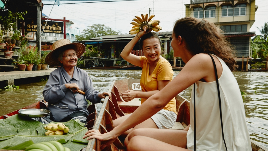 Deux femmes sur un bateau achètent des bananes à une femme qui les vend depuis un autre bateau.
