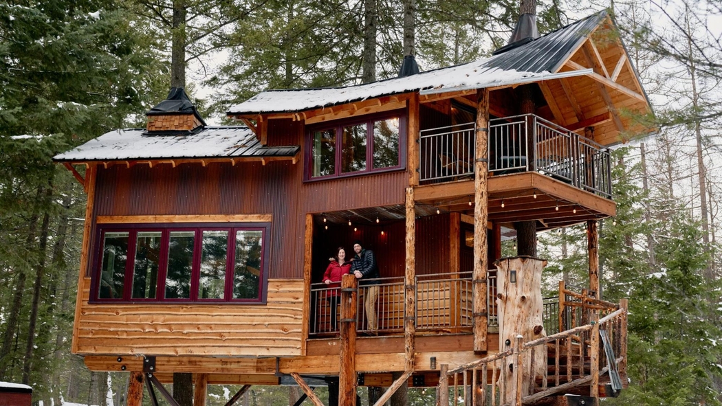 Des hôtes Airbnb se trouvent sur un balcon de leur cabane perchée à Whitefish, dans le Montana. L'extérieur est boisé et le sol enneigé.