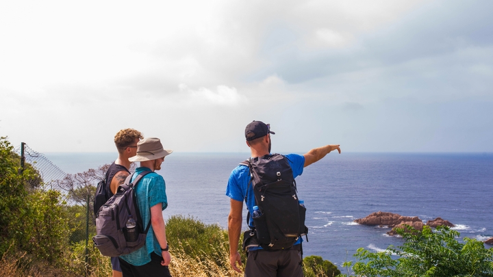 Three people wearing sunglasses, shorts, and backpacks stand on a hill. One of them points out toward the ocean and horizon.