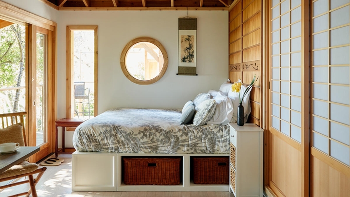 A bedroom with a circular window on the wall and a yellow flower in a vase on the nightstand.