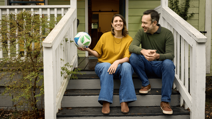 Dve odrasle osobe sede jedna pored druge na stepenicama ispred doma. Oboje se smeše. Jedna drži fudbalsku loptu.