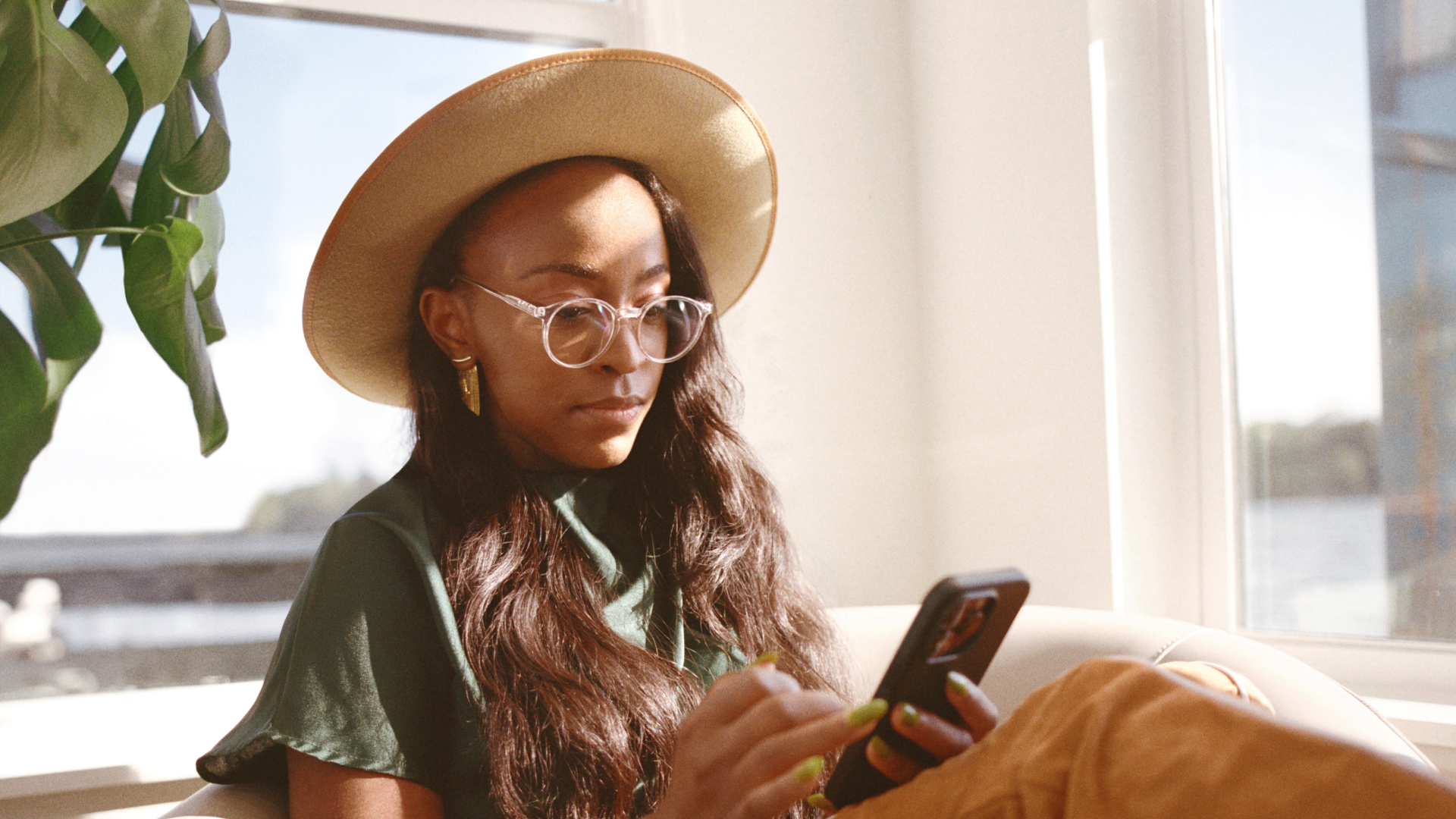 A person wearing glasses and a wide-brimmed hat sits in a beige chair in the sunlight while looking at a smartphone.