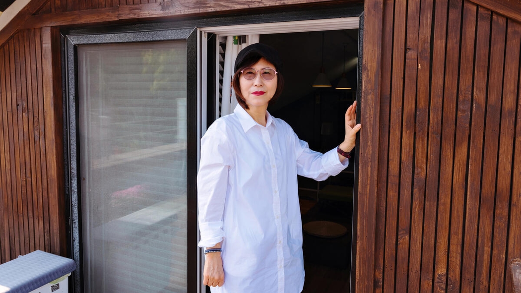 Una persona posa in piedi davanti a una porta-finestra aperta. Con il rossetto rosso, indossa una camicia bianca e occhiali fumé.