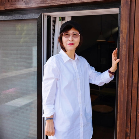 Eine Person steht in einer offenen Tür. Sie trägt ein weißes Hemd mit Knopfleiste, eine getönte Brille und roten Lippenstift.
