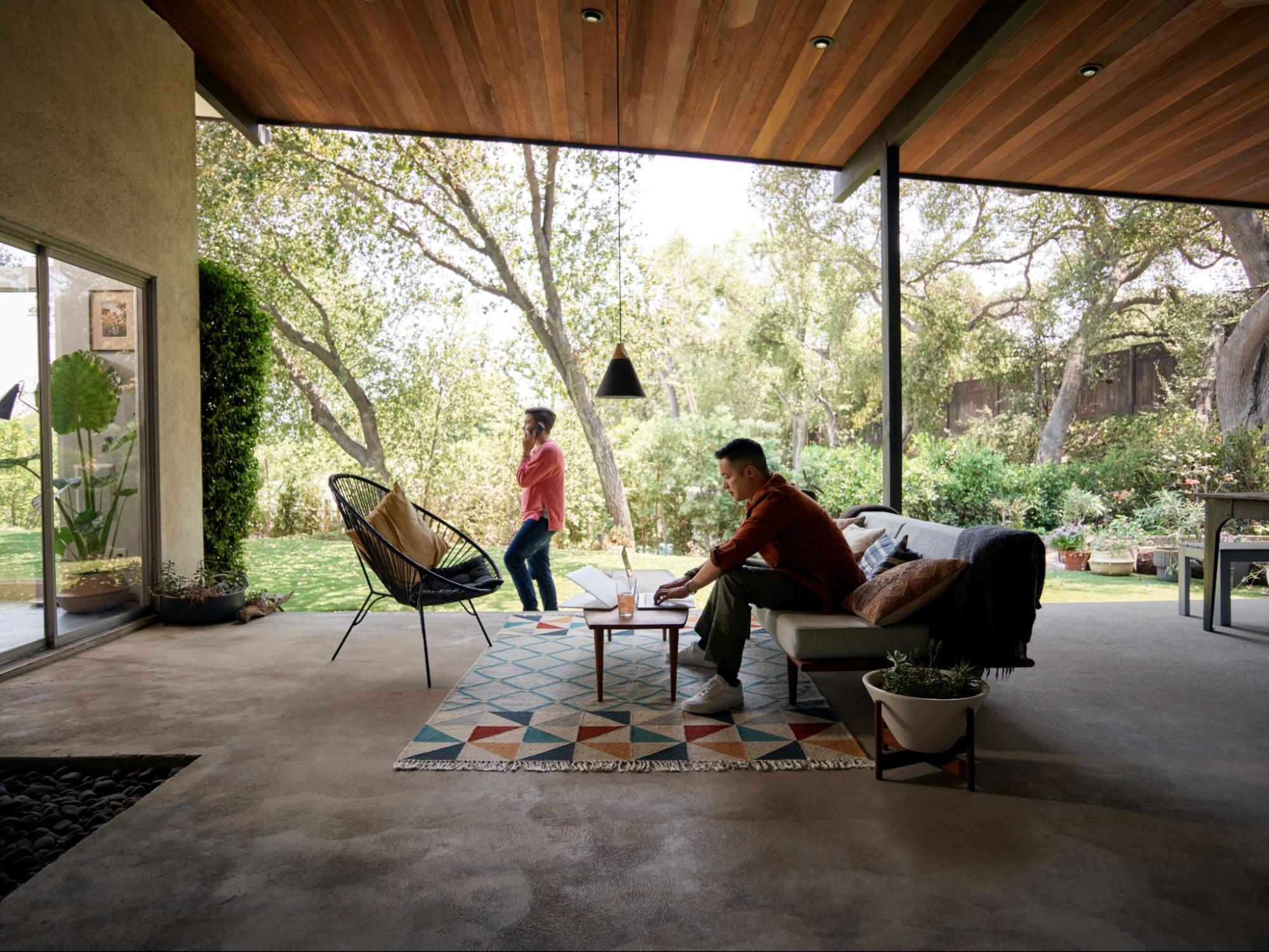 एक व्यक्ति बड़े-से आउटडोर बरामदे में काउच पर बैठकर लैपटॉप पर काम कर रहा है। बैकग्राउंड में एक दूसरा व्यक्ति फ़ोन पर बातें कर रहा है।