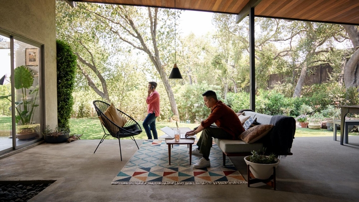 A person sits on a sofa on a large outdoor patio and works on a laptop. Another person talks on the phone in the background.